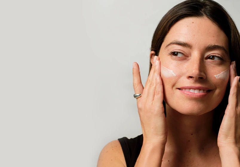 A woman with white cream on her cheeks gently touching her face, shown in a close-up lifestyle shot against a light gray background.