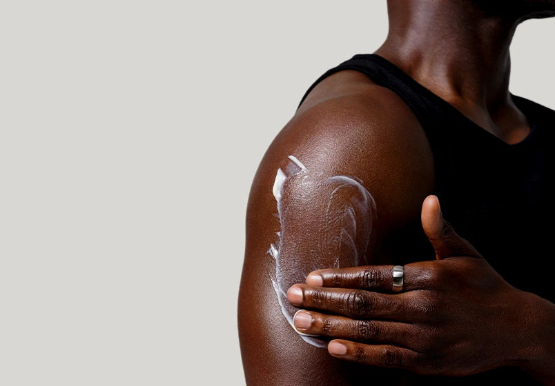 A close-up of a person applying white sunscreen lotion to their upper arm and shoulder while wearing a black tank top, set against a solid light gray background.