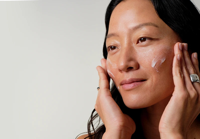 A close-up of a woman with white cream applied to her cheeks, gently touching her face with both hands against a plain light gray background.