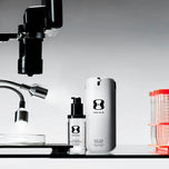 Two ONE SKIN skincare bottles, a smaller black pump bottle and a larger white cylindrical container, displayed on a black surface with a gray gradient background and scientific props including a microscope and petri dish.