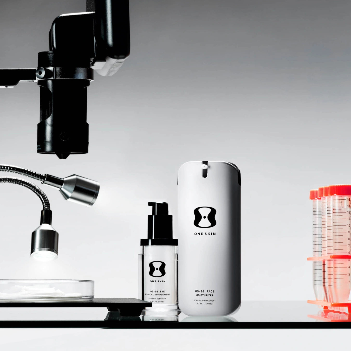 Two ONE SKIN skincare bottles, a smaller black pump bottle and a larger white cylindrical container, displayed on a black surface with a gray gradient background and scientific props including a microscope and petri dish.