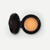 An open black makeup compact showing a smooth, light peachy-beige cream product, photographed on a clean white background.