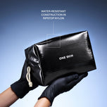 A glossy black zip pouch with white 'ONE SKIN' text, held by hands wearing black gloves against a blue gradient background. White text at the top reads 'WATER-RESISTANT CONSTRUCTION IN RIPSTOP NYLON' with a pointer to a square on the pouch.