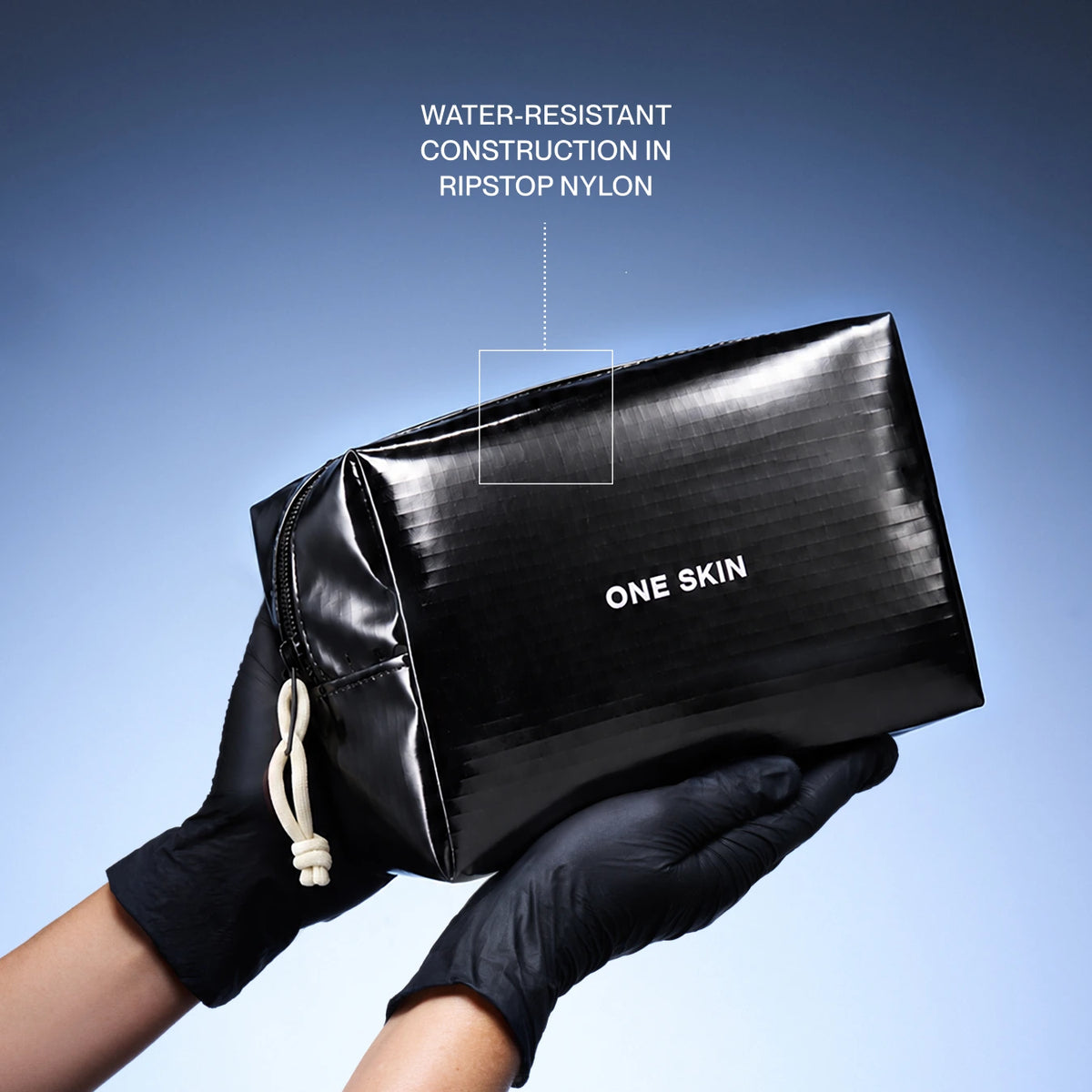 A glossy black zip pouch with white 'ONE SKIN' text, held by hands wearing black gloves against a blue gradient background. White text at the top reads 'WATER-RESISTANT CONSTRUCTION IN RIPSTOP NYLON' with a pointer to a square on the pouch.