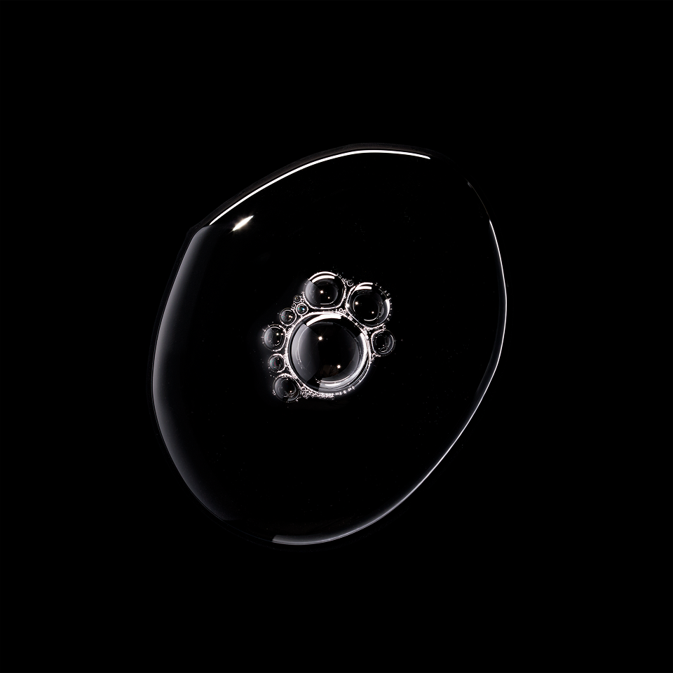A single clear, teardrop-shaped liquid droplet with a cluster of small bubbles in the center, isolated on a solid black background.