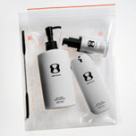 Three skincare products from the brand One Skin, including a large pump bottle, a cylindrical bottle, and a small tube, displayed inside a clear zippered plastic bag with a black spatula tool on a white background.