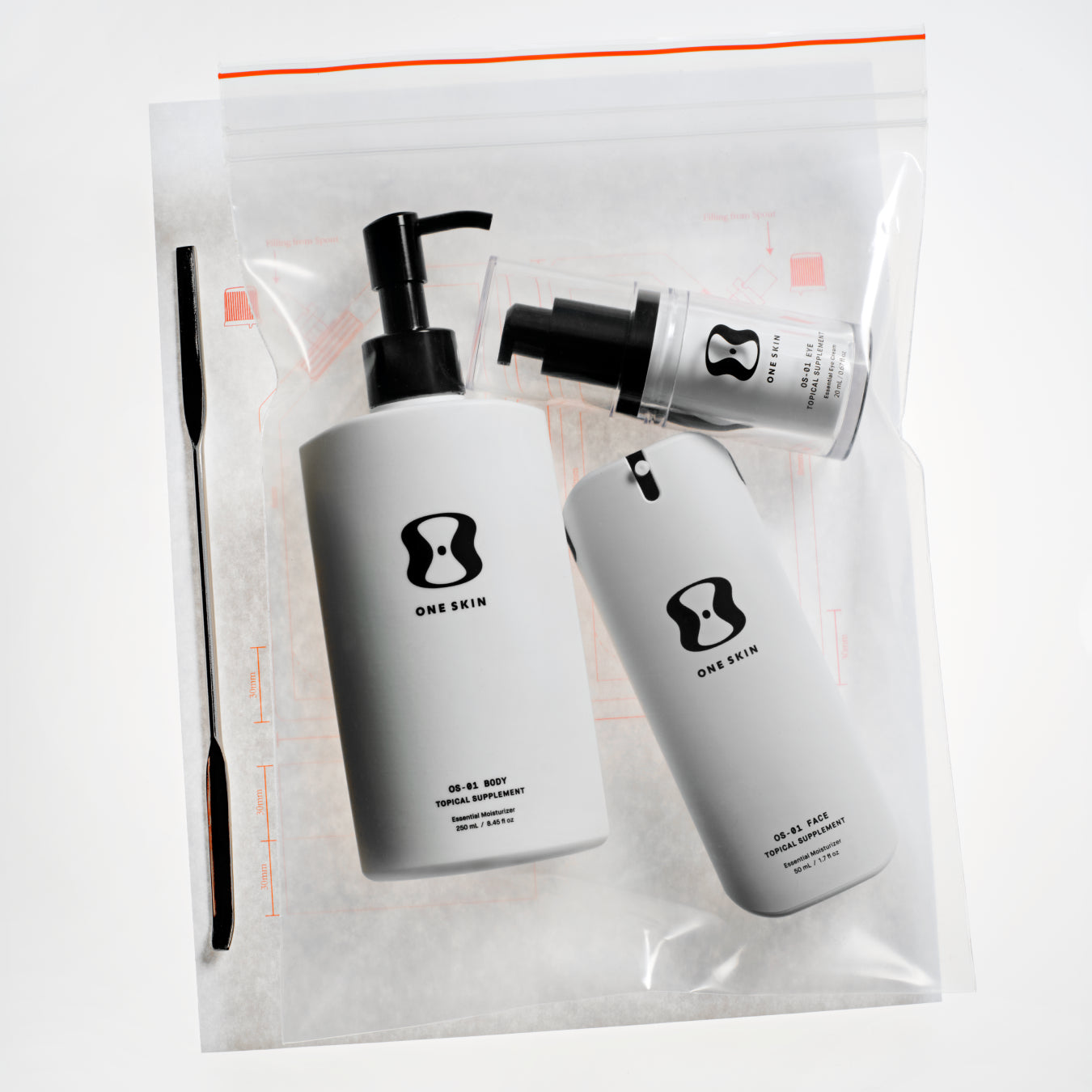 Three skincare products from the brand One Skin, including a large pump bottle, a cylindrical bottle, and a small tube, displayed inside a clear zippered plastic bag with a black spatula tool on a white background.