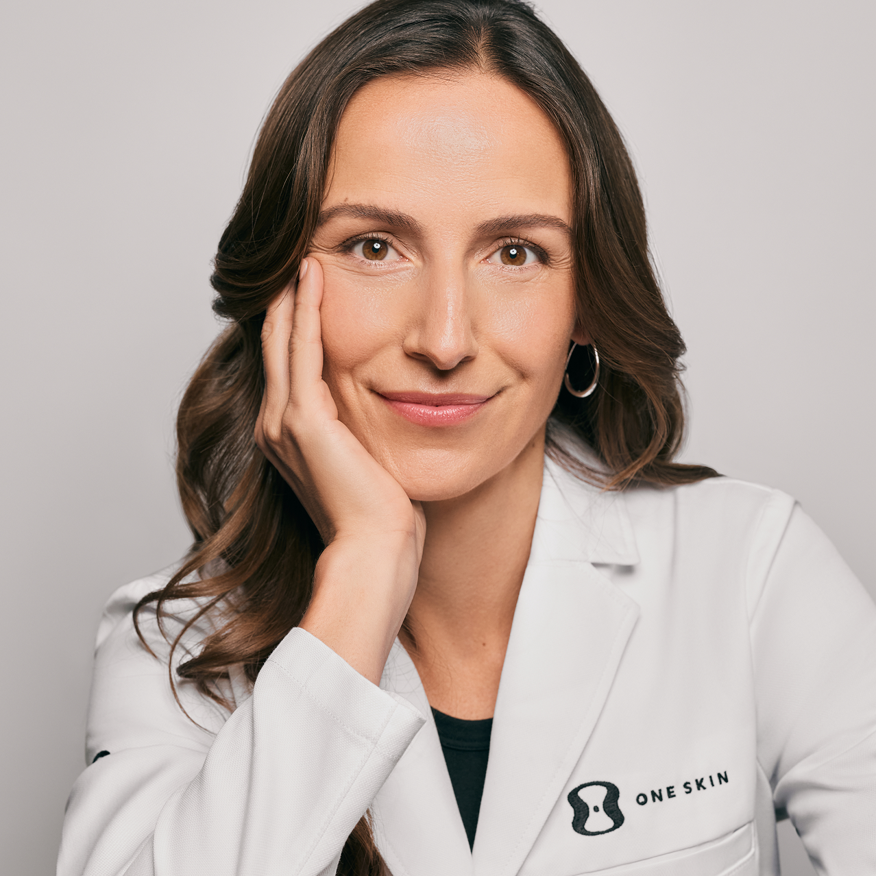 A close-up portrait of a woman with long brown hair wearing a white lab coat with the brand name 'ONE SKIN' embroidered on the left chest, posing with her hand on her cheek against a light gray background.