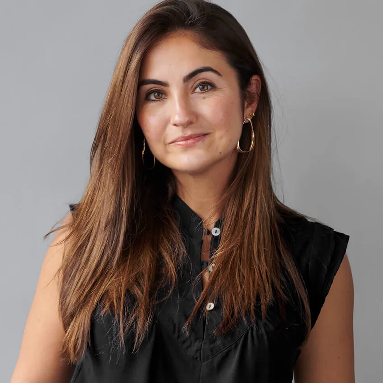 A woman with long brown hair wearing a black sleeveless button-up top and gold hoop earrings, photographed against a plain gray background.