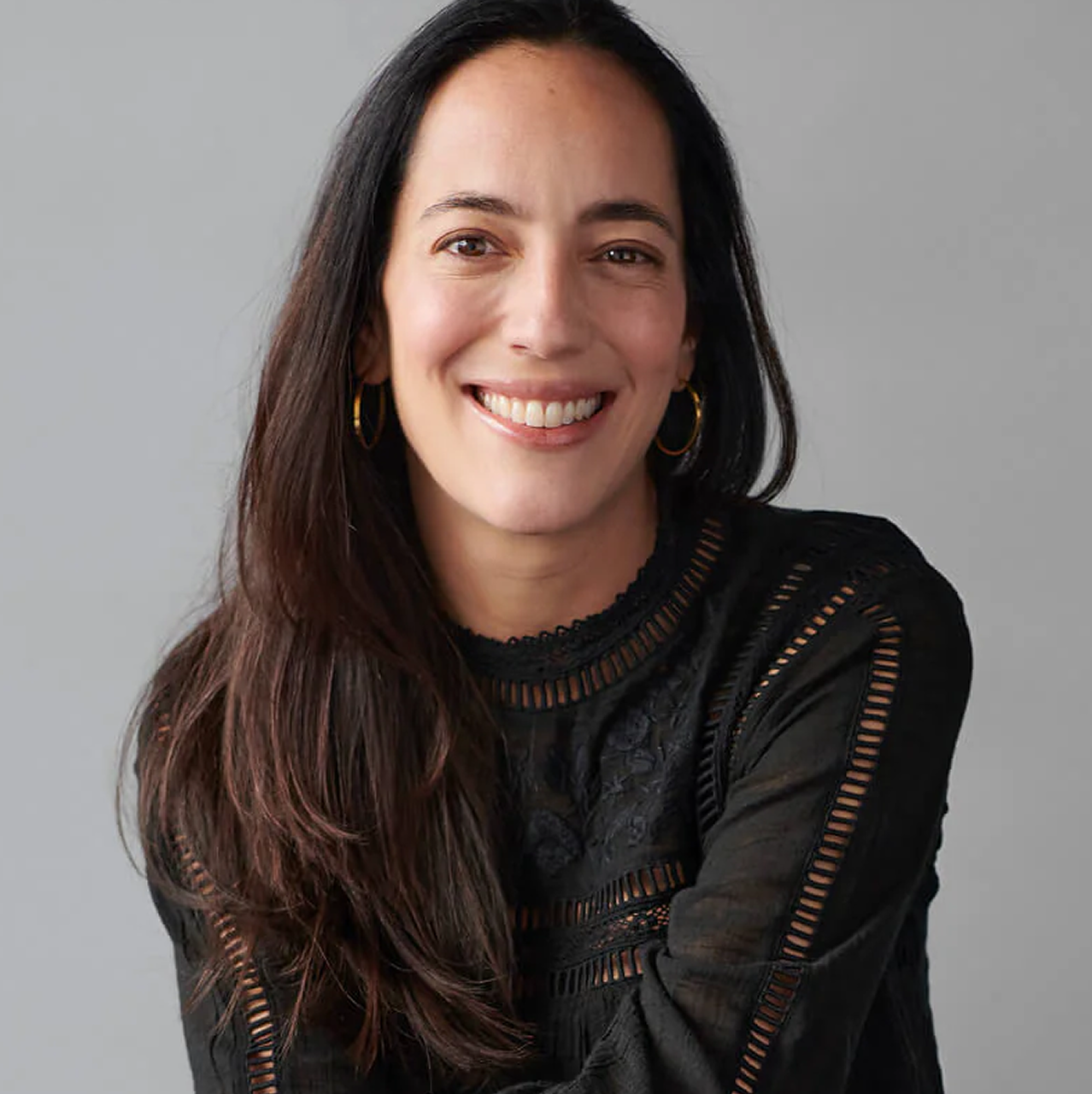 A smiling woman with long dark hair wearing a black long-sleeve top with sheer cutout details and gold hoop earrings, photographed against a plain light gray background.