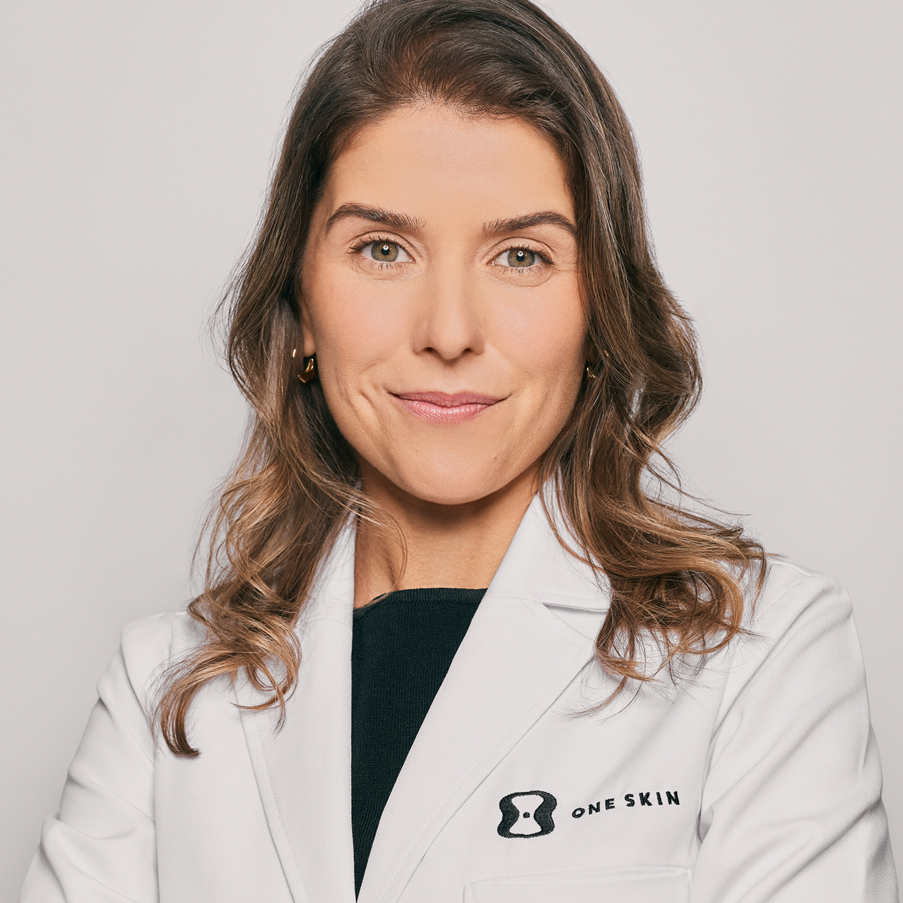 A professional portrait of a woman with brown hair wearing a white lab coat with the 'ONE SKIN' logo on the left chest, set against a solid light gray background.
