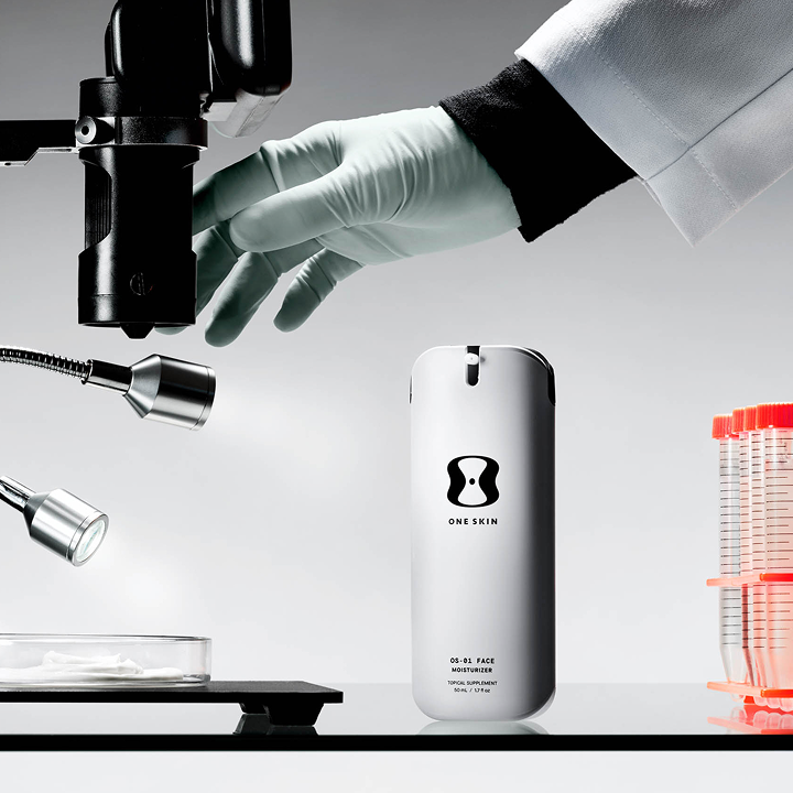 A white cylindrical bottle labeled ONE SKIN stands on a black surface in a clean studio setup. A gloved hand and laboratory tools, including a microscope, ring lights, and stacked test tubes, are arranged around it against a light gray background.