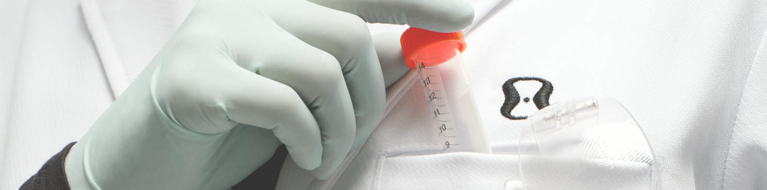 A close-up of a hand in a light green medical glove holding a vial with an orange cap on a white coat.
