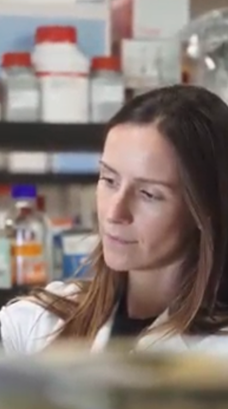 A woman with long brown hair wearing a white lab coat looks to the side in a laboratory setting with shelves of bottles and containers blurred in the background.