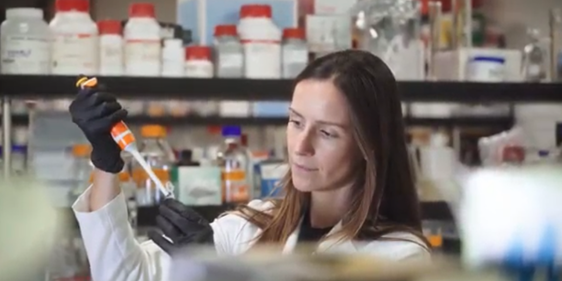 A woman in a white lab coat and black gloves holds a pipette in a laboratory setting with shelves of bottles in the background.