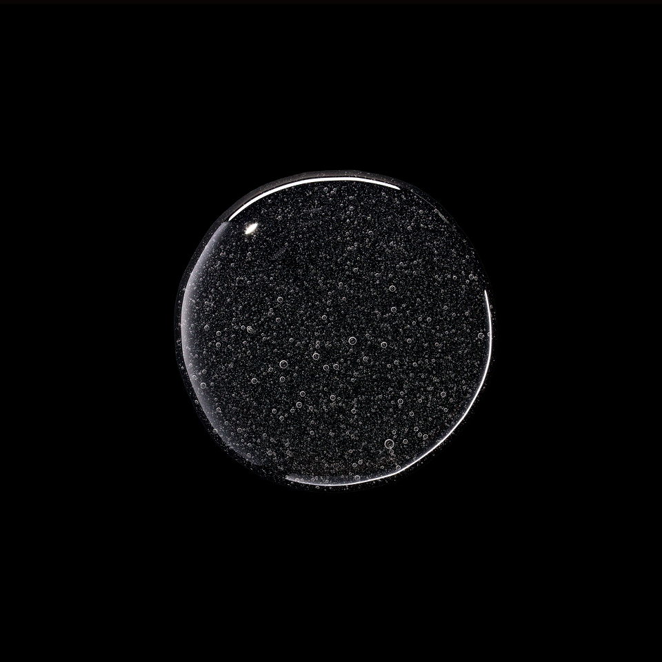 A clear, circular cosmetic or material swatch showing a dense texture of tiny black and white speckles, isolated on a solid black background.
