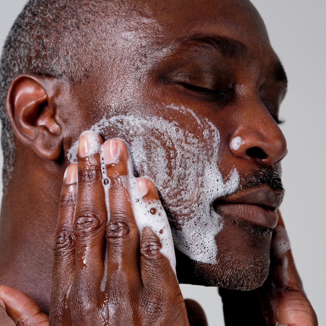 Close-up of a man washing his face with foamy soap, showing his cheek, hand, ear, and part of his nose against a light gray background.